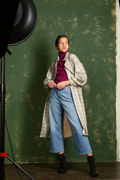 teen girl in checkered trench coat, jeans and bootswalking full body portrait on green wall background - 写真・画像