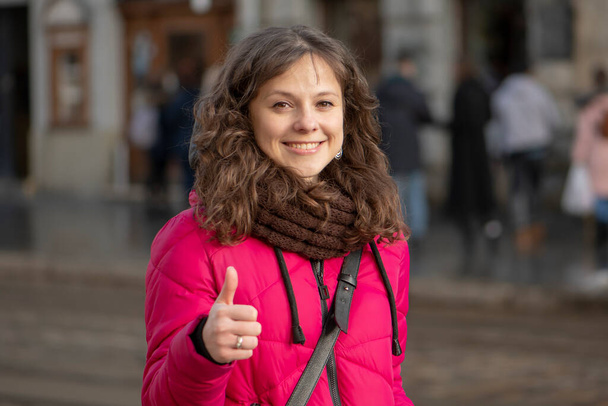 Street portrait of a cheerful woman 30-35 years old with curly hair and delicate features on the background of the urban landscape. The woman holds her thumbs up. Concept: everything is fine ! - Photo, Image