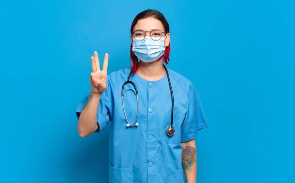 young attractive red hair woman smiling and looking friendly, showing number three or third with hand forward, counting down. hospital nurse concept - Foto, imagen