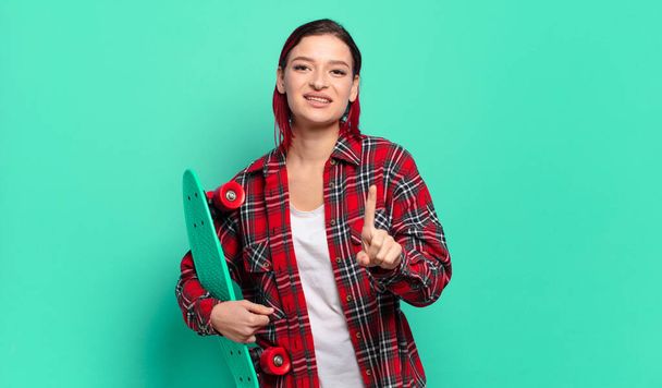 young attractive red hair woman smiling proudly and confidently making number one pose triumphantly, feeling like a leader and holding a skate board - Foto, afbeelding