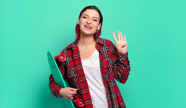 young attractive red hair woman smiling and looking friendly, showing number four or fourth with hand forward, counting down and holding a skate board - Photo, image