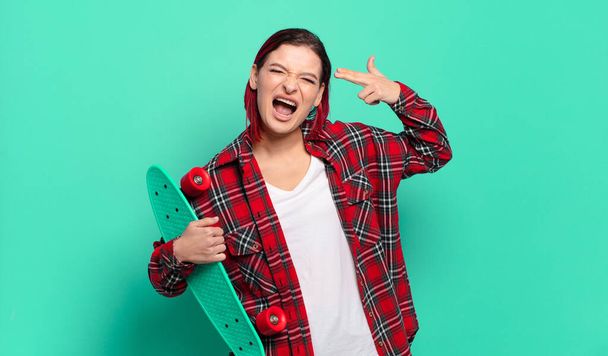 young attractive red hair woman looking unhappy and stressed, suicide gesture making gun sign with hand, pointing to head and holding a skate board - Foto, immagini