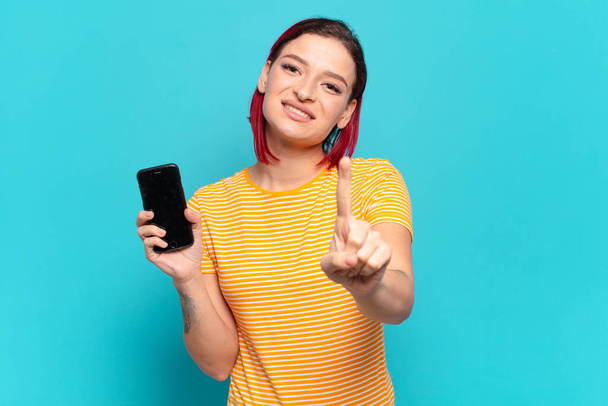 young attractive red hair woman smiling proudly and confidently making number one pose triumphantly, feeling like a leader and showing her cell - Fotografie, Obrázek