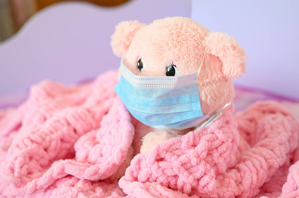Um ursinho de pelúcia rosa em uma máscara médica e um termômetro fica coberto com um cobertor rosa. Conceito de coronovírus ou gripe. Focando no olho do urso. - Foto, Imagem