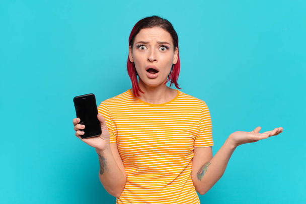 young attractive red hair woman open-mouthed and amazed, shocked and astonished with an unbelievable surprise and showing her cell - Foto, imagen