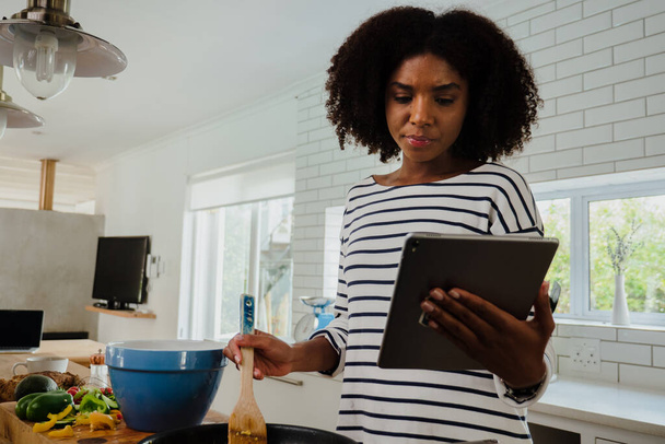 Schöne Mischlingsfrau schaut sich Rezept auf Tablet an, während sie zu Hause in ihrer Küche kocht - Foto, Bild