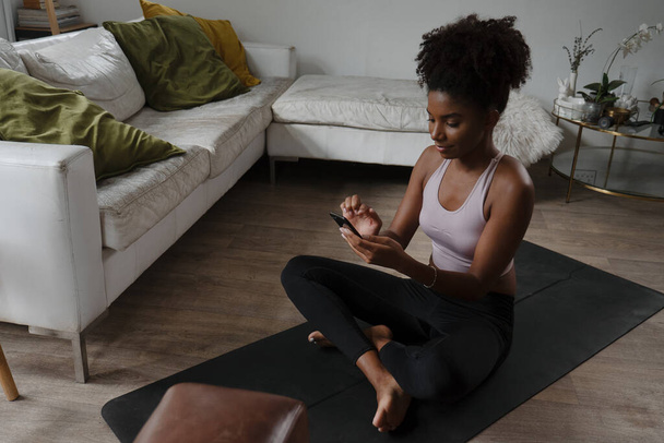 African American female teen choosing yoga flow on smartphone getting ready to exercise in modern lounge - Photo, Image