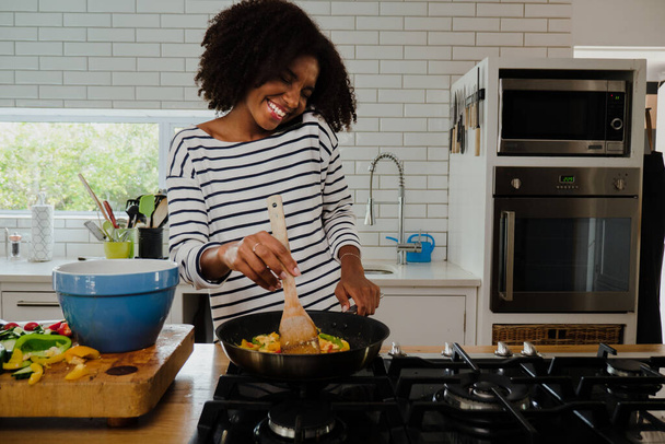 Uśmiechnięta Afroamerykanka robi pyszną kolację rozmawiając z chłopakiem na smartfonie przygotowując się na randkę w fantazyjnej kuchni. - Zdjęcie, obraz