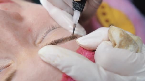 Uma máquina de tatuagem de agulha especial faz correção permanente de maquiagem das sobrancelhas de uma jovem. Pigmento escuro injetado sob a pele. Microblading, pulverização de pó close-up. - Filmagem, Vídeo