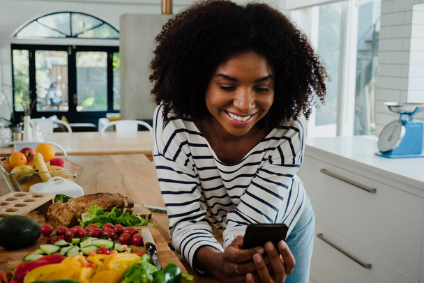 Kaunis nainen afro keittiössä selaamassa mobiililaitteella - Valokuva, kuva