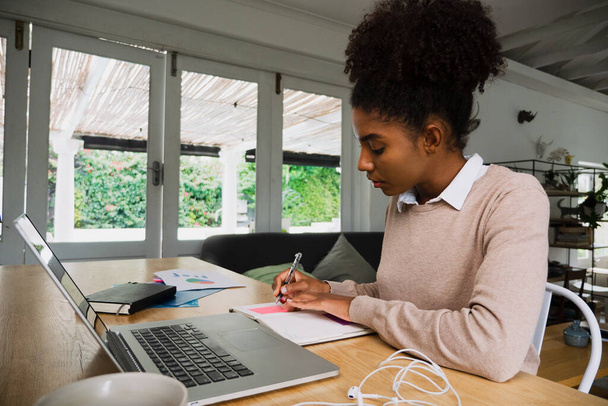 Sekarotuinen nainen opiskelija kirjoittaa muistiinpanoja katsellessaan verkossa luento kannettavan istuu iso työpöytä keittiössä - Valokuva, kuva