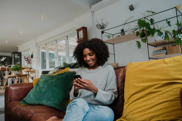 Genç, melez, afro saçlı bir kadın evdeki kanepede akıllı telefondan sohbet ediyor. - Fotoğraf, Görsel