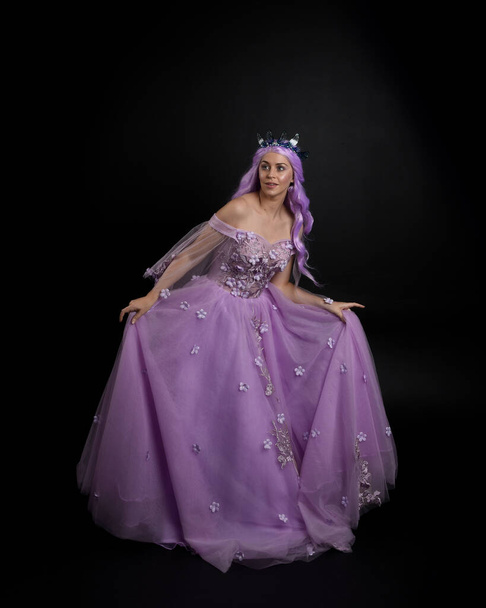 Retrato de longitud completa de la niña con vestido largo de bola de fantasía púrpura con corona y pelo rosa, pose de pie con movimientos gestuales elegantes contra un fondo de estudio. - Foto, imagen