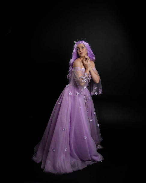 Teljes hosszúságú portré a lány visel hosszú lila fantasy labda ruha korona és rózsaszín haj, álló póz elegáns geszturális mozgások ellen stúdió háttér. - Fotó, kép
