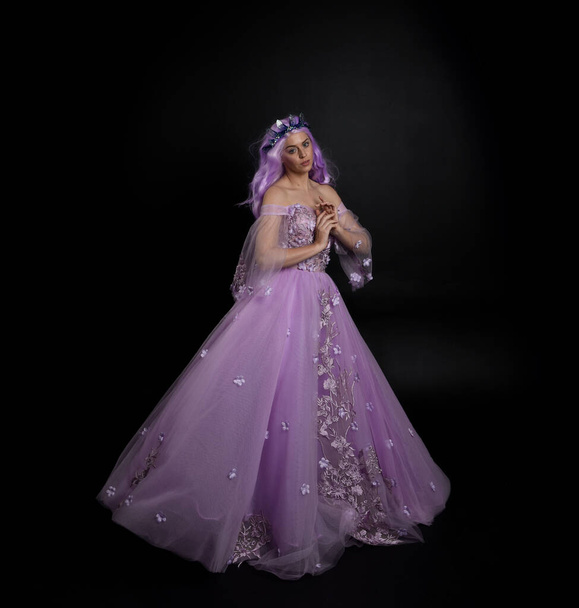 Full length portrait of girl wearing long purple fantasy ball gown with crown and pink hair, standing pose with elegant gestural  movements against a studio background. - Photo, Image