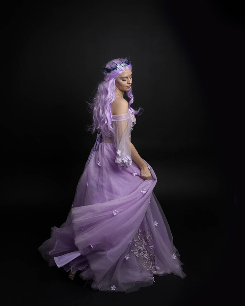 Ganzkörperporträt eines Mädchens in einem langen lila Fantasie-Ballkleid mit Krone und rosa Haaren, das mit dem Rücken zur Kamera vor Studiohintergrund posiert. - Foto, Bild