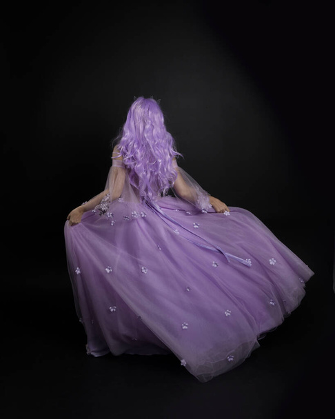 Full length πορτρέτο του κοριτσιού φορώντας μακρύ μωβ φόρεμα μπάλα φαντασίας με στέμμα και ροζ μαλλιά, στέκεται ποζάρουν με την πλάτη στην κάμερα σε φόντο στούντιο. - Φωτογραφία, εικόνα