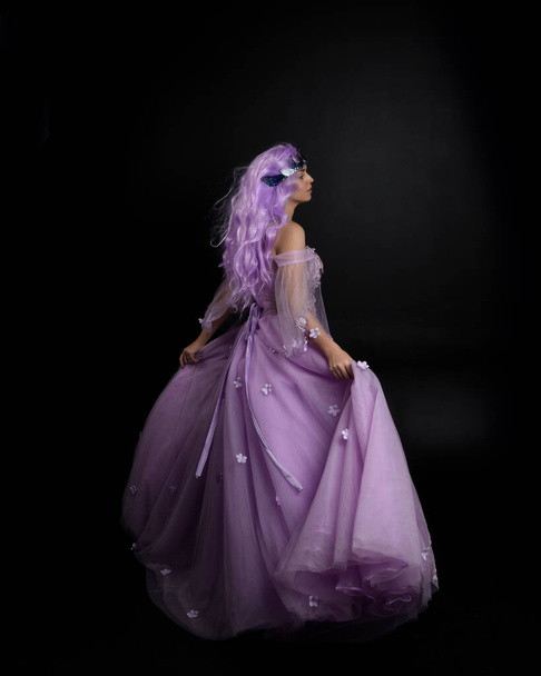 Full length portrait of girl wearing long purple fantasy ball gown with crown and pink hair, standing pose with back to the camera  against a studio background. - Photo, Image