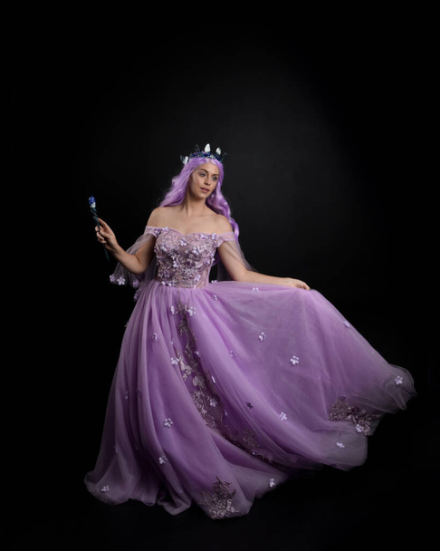 Portrait pleine longueur de fille portant une longue robe de bal fantaisie pourpre avec couronne et cheveux roses, pose debout tenant une baguette sur un fond de studio. - Photo, image