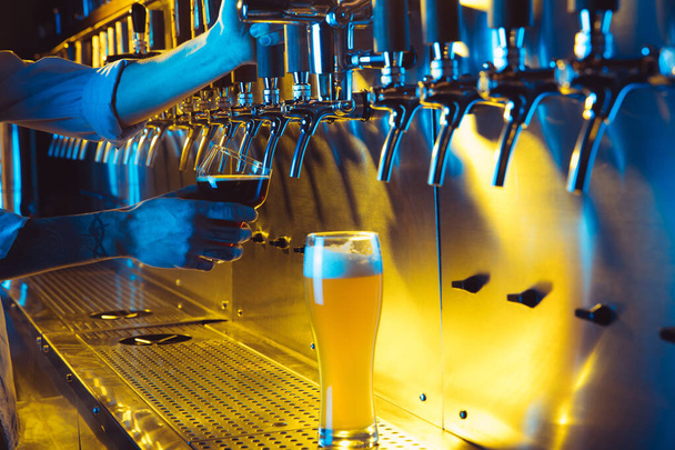 Baarimikon käsi kaataa tuoretta tummaa olutta hanasta lasiin neonvalossa. - Valokuva, kuva