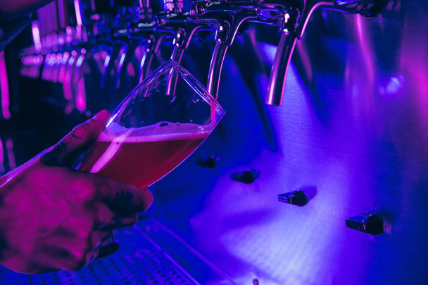 Γκρο πλαν ποτήρι μπύρα στο χέρι μπάρμαν σε πολύχρωμο νέον φως. - Φωτογραφία, εικόνα