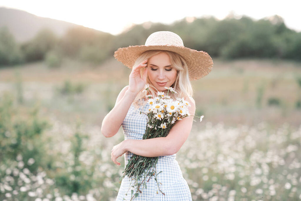 美しいブロンドの女性を笑顔25-29歳の新鮮なデイジーの花束を保持しているわらの帽子を身に着けており、牧草地でポーズをとるドレスを閉じます。夏時間だ。幸せよ。健康的なライフスタイル.  - 写真・画像