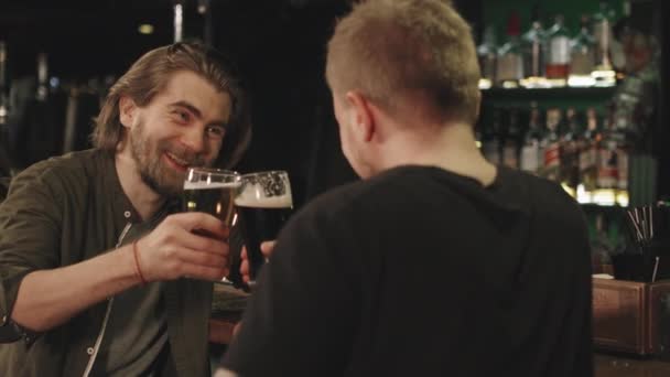 Slow-motion waist-up shot of joyful young men chilling at bar in evening, toasting beer mugs while talking - Footage, Video