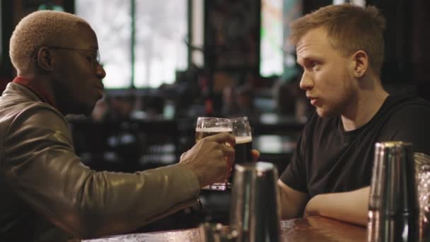 Seitliche Aufnahme mit langsamen, multiethnischen jungen Männern, die sich unterhalten, während sie abends in einem lokalen Irish Pub Bier trinken - Filmmaterial, Video