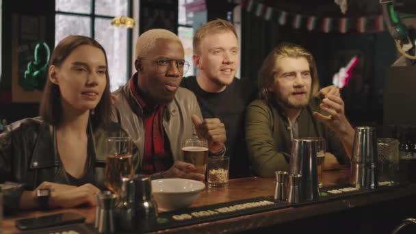 Waist-up shot of joyful multi-ethnic friends sitting at bar in evening watching sports game on TV, cheering emotionally after goal - Footage, Video