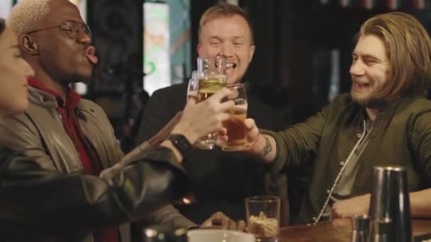 Plan moyen au ralenti d'une jeune compagnie joyeuse d'amis multiethniques s'amusant ensemble au pub local, grillant des verres à bière et riant - Séquence, vidéo