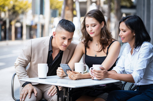 Группа молодых бизнесменов, сидящих в городском уличном кафе, разговаривает и наслаждается. Они обсуждают что-то на смартфоне.. - Фото, изображение