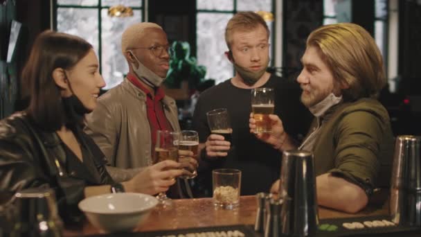 Mediana toma de cuatro amigos multiétnicos que se divierten juntos bebiendo cerveza en el pub local por la noche, haciendo tostadas y tazas de cerveza tintineo - Imágenes, Vídeo