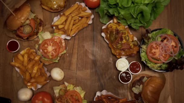 Servir un hamburger sur une table pleine de repas de restauration rapide. Vue aérienne du haut vers le bas des aliments frits sur une table en bois - Séquence, vidéo