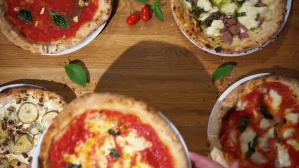 伝統的なイタリアのピザにバジル、チーズ、トマトソースを木製のテーブルで提供しています。テーブルの上から様々なおいしいピザの景色を見下ろす - 映像、動画