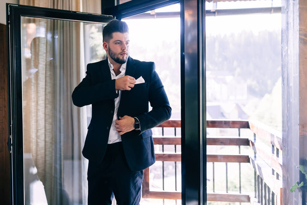 Groom's morning before the wedding. Morning preparation. A young and handsome man is dressing in a wedding suit by the window. - Foto, imagen