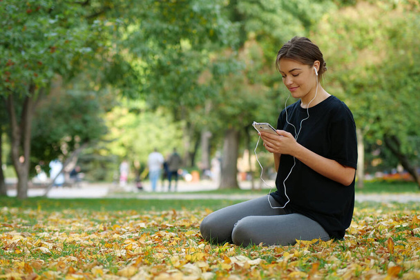 Junges schönes Mädchen sitzt auf gelbem Laub auf Rasen im herbstlichen Stadtpark, nutzt Smartphone und hört Musik - Foto, Bild