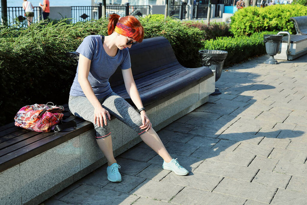 Young red-haired woman in sportswear sits on bench outdoors and touches her sore knee on sunny summer day - Photo, Image