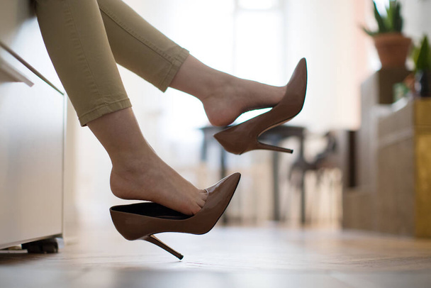 Yorgun kadın işten sonra kahverengi topuklu ayakkabılarını çıkararak dinleniyor ya da yürüyor, kanepede uzanıyor. rahatsız ayakkabılar - Fotoğraf, Görsel