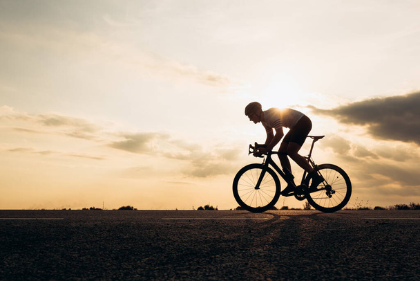 Widok z boku aktywnego sportowca w kasku ochronnym jazda na rowerze wśród przyrody podczas zachodu słońca. Muskularny facet w stroju aktywnym jeżdżący po utwardzonej drodze. - Zdjęcie, obraz