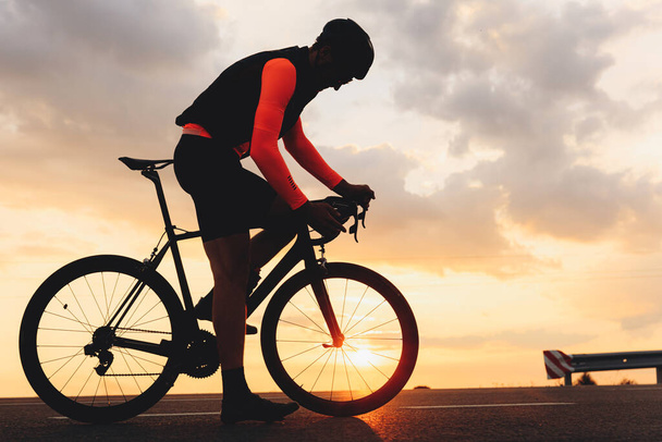 Vista laterale del ciclista muscoloso vestito con abiti sportivi luminosi in piedi su strada con la sua bici professionale per le corse. Incredibile tramonto su sfondo. - Foto, immagini
