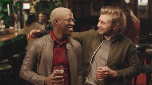 Retrato mediano en cámara lenta de dos alegres amigos varones multiétnicos con vasos de cerveza en las manos mirando a la cámara sonriendo de fiesta en un pub local juntos - Imágenes, Vídeo