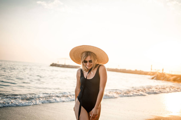 Молодая красивая женщина в черном купальнике и шляпе в очках прогуливается по пляжу Турции на закате. Концепция морского отдыха. Селективный фокус - Фото, изображение