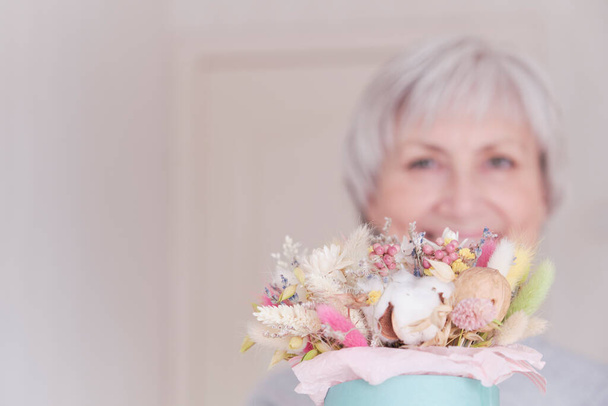 Ένα πορτραίτο μιας ηλικιωμένης Καυκάσιας γυναίκας με κοντά γκρίζα μαλλιά και ένα μπουκέτο αποξηραμένα λουλούδια. Επιλεκτική εστίαση στην ανθοδέσμη. - Φωτογραφία, εικόνα