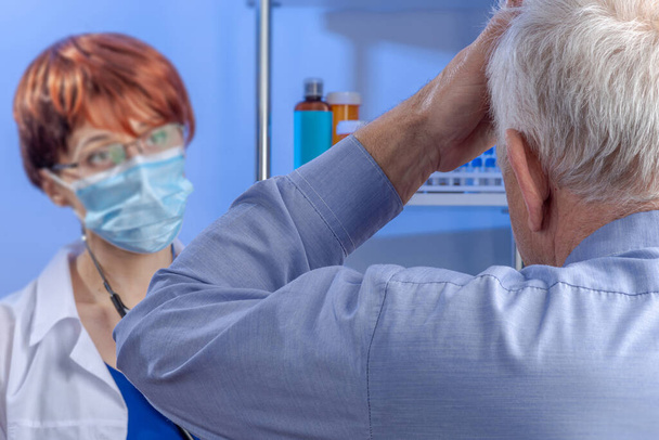 Un paciente de pelo gris mayor apoyando su mano sobre su cabeza quejándose a un joven doctor de una migraña. Atención médica profesional en el hospital. - Foto, imagen