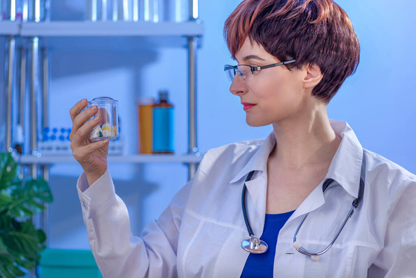 Doctora joven con el pelo corto con estilo en blanco abrigo médico sosteniendo frasco de píldoras en la clínica. - Foto, imagen