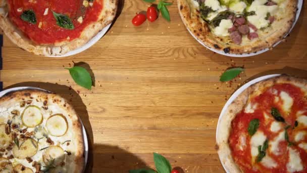 木製のテーブルの上にバジル、チーズ、トマトソースとイタリアのピザ。レストランのテーブルで提供される様々なピザの品揃えのトップダウンビュー - 映像、動画