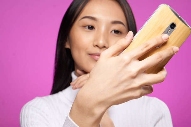 hezká asijská žena s telefonem v ruce při pohledu na obrazovce close-up růžové pozadí - Fotografie, Obrázek