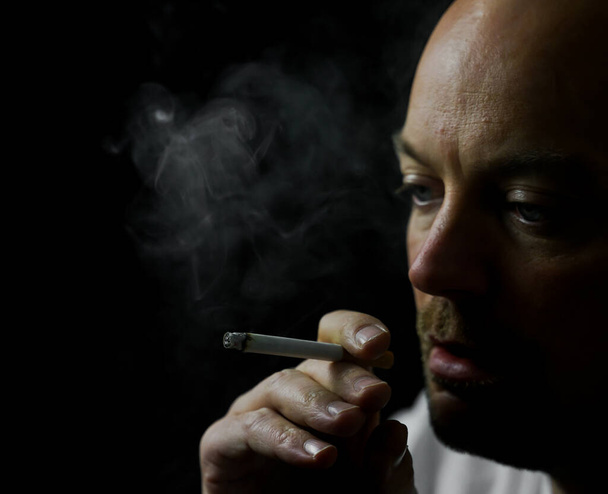 Sigara içen bir adam beyaz duman üflüyor. Siyah arkaplanda izole edilmiş, alan derinliği sığ. Yüksek kalite fotoğraf - Fotoğraf, Görsel