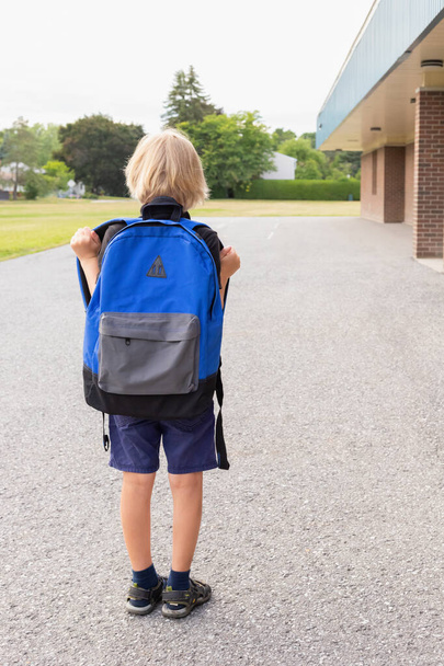 Ένα μικρό ξανθό παιδί κουβαλάει μπλε σακίδιο, πηγαίνει στο σχολείο. Επιστροφή στο σχολείο έννοια. Παιδί κοντά στο κτίριο του σχολείου στην αυλή του σχολείου. - Φωτογραφία, εικόνα