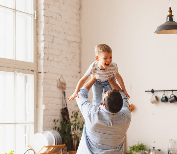 Faceless carefree father in casual clothes lifting adorable little son above head while having fun together in contemporary kitchen - Photo, Image